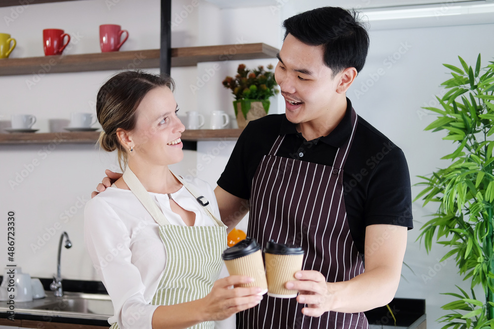 Smiling young barista couple loves Asian man and caucasian woman holding cups of hot coffee together. Start up Coffee shop and cafe business concept Smiling young barista couple loves Asian man and caucasian woman holding cups of hot coffee together. Start up Coffee shop and cafe business concept