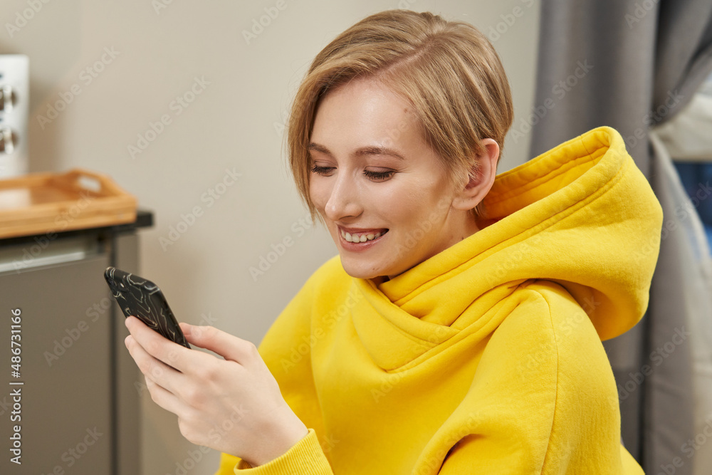 beautiful young woman with a short haircut in a yellow sweatshirt looks at something on the phone and smiles beautiful young woman with a short haircut in a yellow sweatshirt looks at something on the phone and smiles