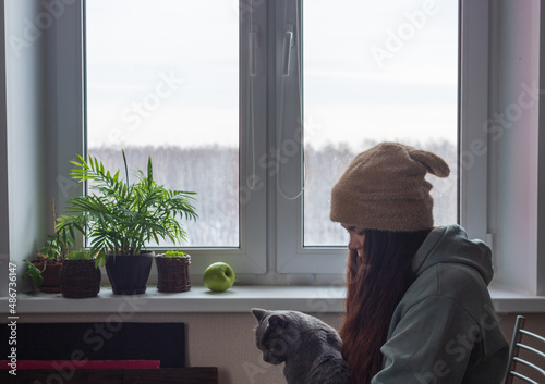 A woman in a hat with ears and long hair is sitting by the window with her cat