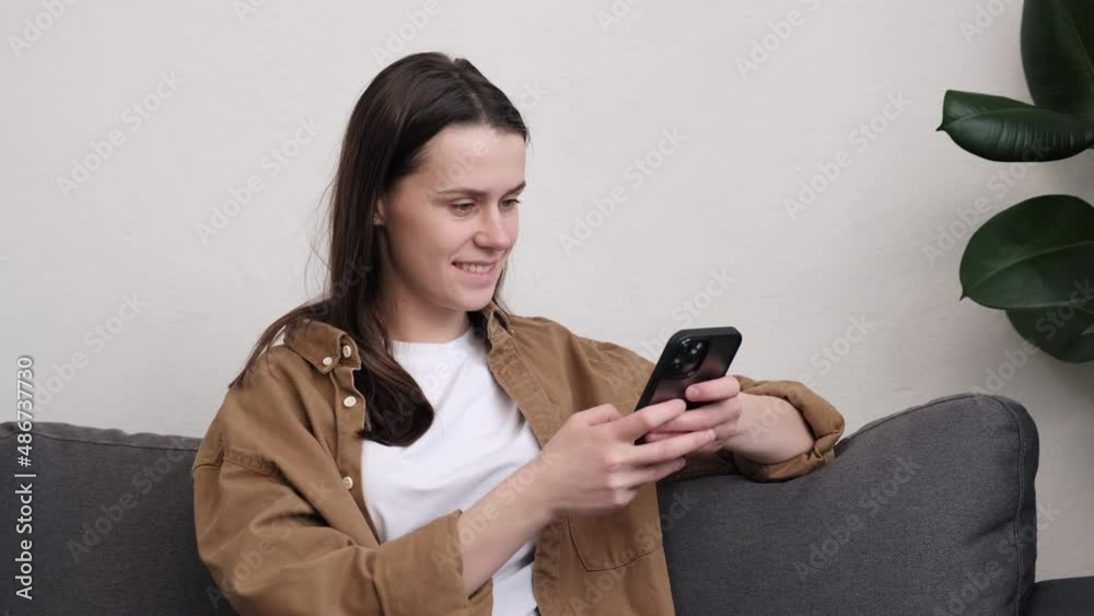 Close up of smiling happy young woman holding cell phone sitting on cozy sofa at home, using online mobile apps technology doing online shopping, dating in application, checking social media, texting Close up of smiling happy young woman holding cell phone sitting on cozy sofa at home, using online mobile apps technology doing online shopping, dating in application, checking social media, texting