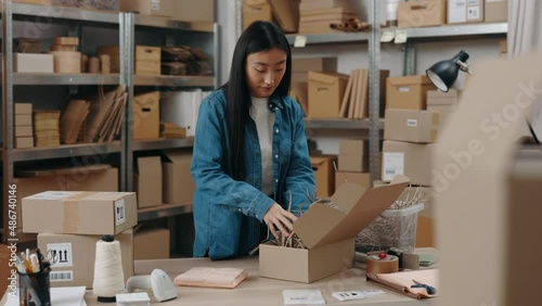 Waist up portrait view of the asian woman standing at the warehouse and putting filling at the cupboard box while packing parcels at the wooden table. Small business concept.