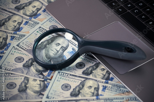 Laptop and magnifying glass on us dollar bills, close up Benjamin Franklin portrait, make money online concept