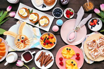 Fun Easter breakfast or brunch table scene. Top down view on a dark wood background. Bunny pancake, egg nests, chick fruit and assorted spring food items.