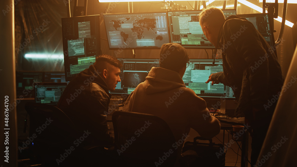 Young man in hoodie helping associate to hack database for criminal while gathering around table in dim room