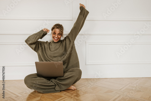 Fotografie Smiling young caucasian woman using laptop while sitting on floor at home
