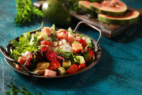 Tropical salad on a bright turquoise background. Fresh salad in a gray plate of arugula  papaya  avocado  cherry tomatoes  strachatella and pine nuts. Exotic dish. Background image  copy space