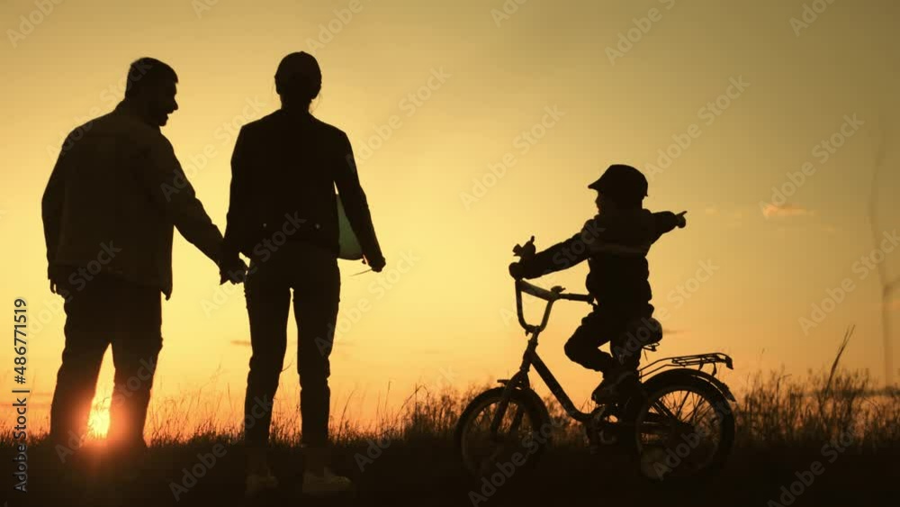People silhouettes on summer sunset meadow. The kid is learning to ride a bike. Time to be together.Silhouette of happy family on a beautiful sunset meadow. People silhouettes on summer sunset meadow. The kid is learning to ride a bike. Time to be together.Silhouette of happy family on a beautiful sunset meadow.