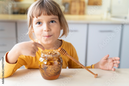 Child girl eating golden honeycomb and beeswax in a jar of honey on the table. Fresh organic honey in a jar with a wooden stick closeup. Background for beekeepers, healthy nutrition organic food