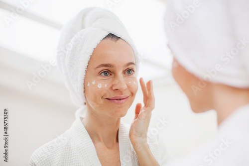 Attractive adult 40s woman apply facial cream middle-aged female look in mirror
