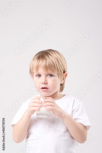 Portrait of a blond child with a glass of milk on a white background. Expressive face, smile, eye contact. Vitamins, calcium, milk teeth, dairy products, healthy food, advertising concept. Copy space