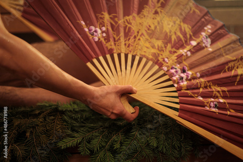 A woman performs a healing ritual with a Chinese fan for a man. A man lies on spruce branches.