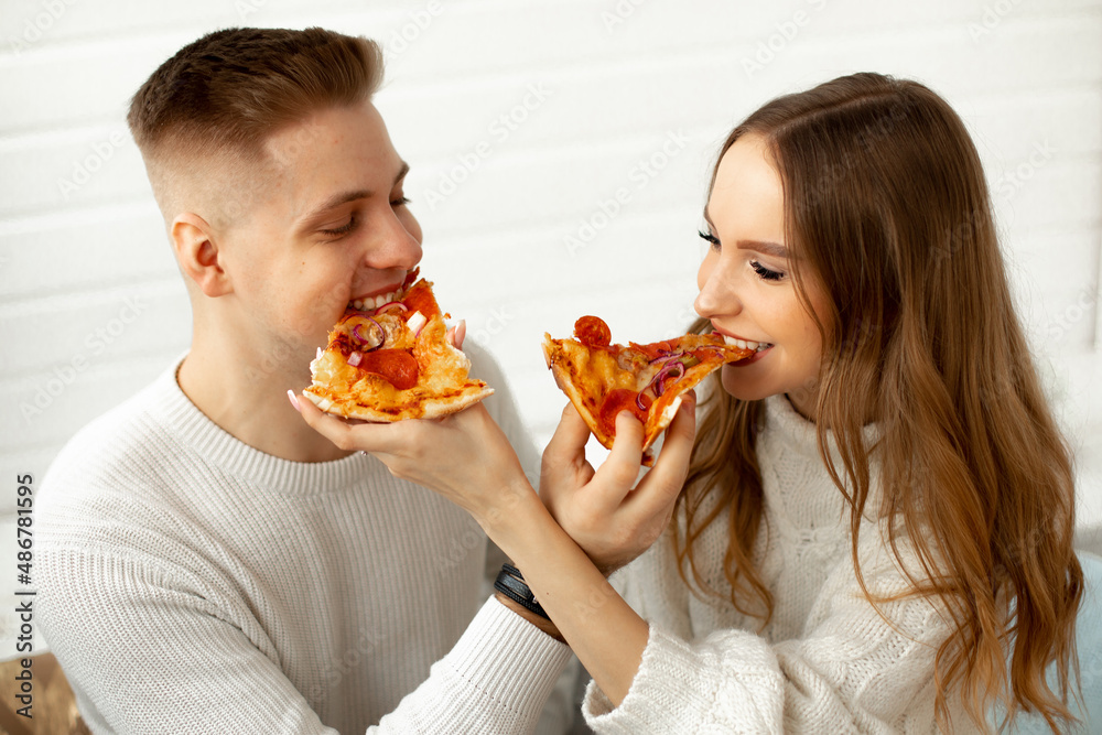 Enjoy pizza.Young woman and her lover are happy, having lunch together and jokingly feeding each other, they are sitting against white wall. Date at home. Romantic relationships. 