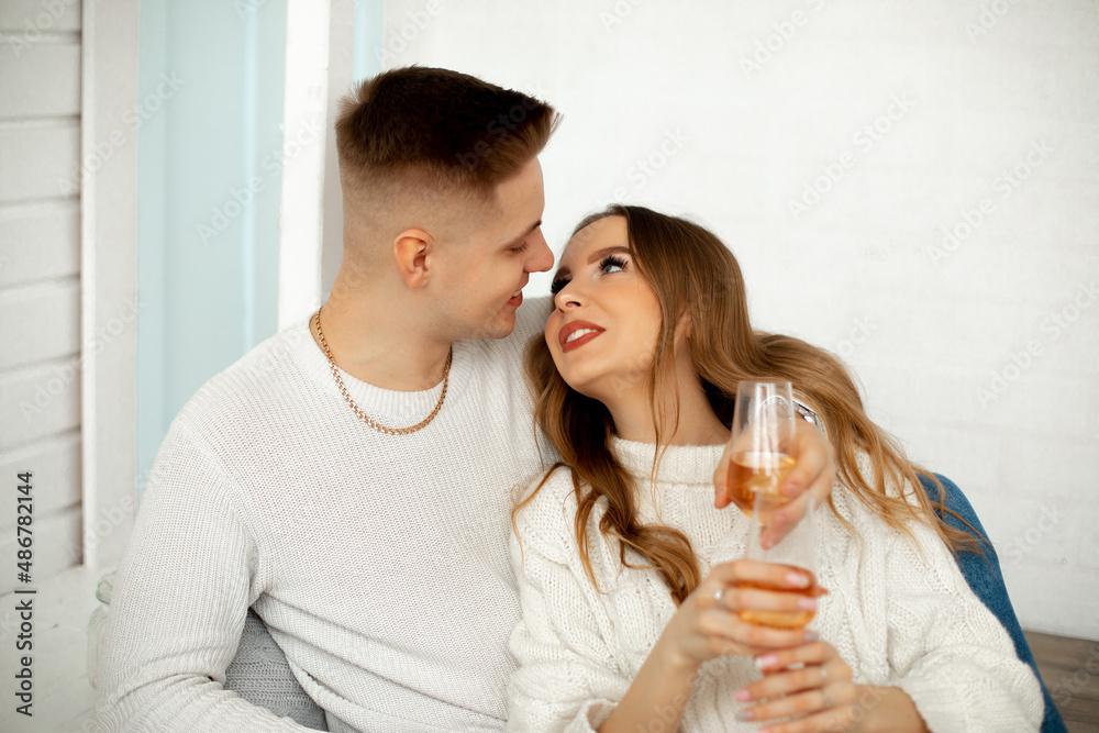 Young girl with brown hair looks at her lover, pressing her back to him, he hugs her shoulders. They are sitting in room with glasses of wine. Romantic. Love and tenderness. Young girl with brown hair looks at her lover, pressing her back to him, he hugs her shoulders. They are sitting in room with glasses of wine. Romantic. Love and tenderness.