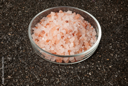 Rose salt in a Petri dish on a ruberoid mineral surface. Black granular background. Pink salty crystals in chemical flat glassware. Natural sea salt granules of chlorides. Himalayan spice. photo