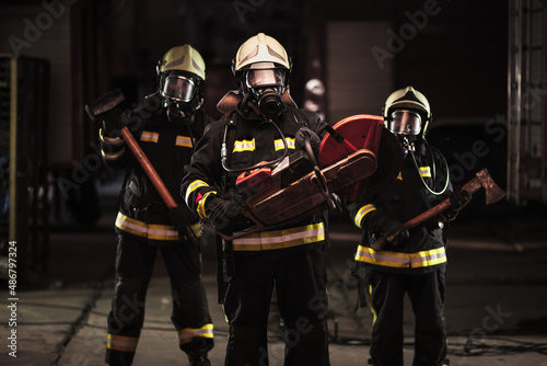 Group of professional firefighters wearing full equipment, oxygen masks, and emergency rescue tools, circular hydraulic and gas saw, axe, and sledge hammer. Firetrucks in the background.