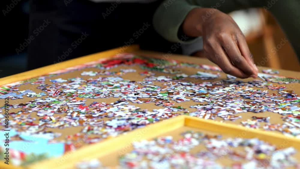 African American woman solving a jigsaw puzzle moves hand over the table and sets down a piece. Hand held closeup clip, with shallow depth of field focus. African American woman solving a jigsaw puzzle moves hand over the table and sets down a piece. Hand held closeup clip, with shallow depth of field focus.
