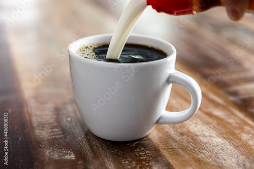фотографія pouring creamer into black coffee in a white mug on a wood table.