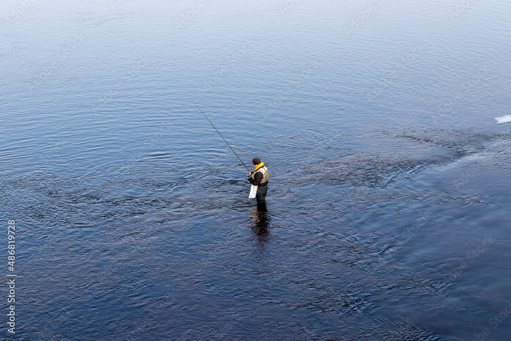 A fisherman stands knee-deep in water with a fishing rod, winter fishing on the river A fisherman stands knee-deep in water with a fishing rod, winter fishing on the river
