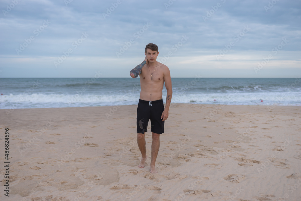 Full height portrait of handsome fit bare-chested young man standing on the beach. guy with tattoo walking by the sea. High quality photo