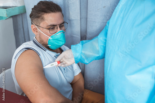 A nurse in PPE gear inserts a digital thermometer in the armpit of a man wearing a N95 mask. Measuring an underarm or axillary temperature. At the emergency ward of a hospital.
