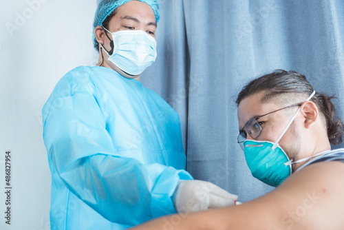 A male nurse in PPE gear inserts a digital thermometer in the armpit of a man wearing a N95 mask. Measuring an underarm or axillary temperature. At the emergency ward of a hospital.