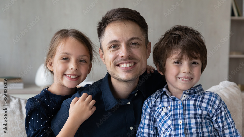 Happy dad and two little sibling kids sitting on sofa at home, looking at camera with toothy smiles. Young father and children posing for family picture together. Head shot portrait, banner Happy dad and two little sibling kids sitting on sofa at home, looking at camera with toothy smiles. Young father and children posing for family picture together. Head shot portrait, banner