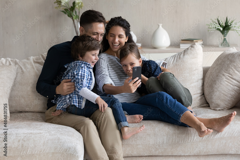 Cheerful family couple and excited happy children resting on sofa, using digital device, taking selfie on smartphone, making video call, looking at mobile phone screen, smiling, laughing, having fun