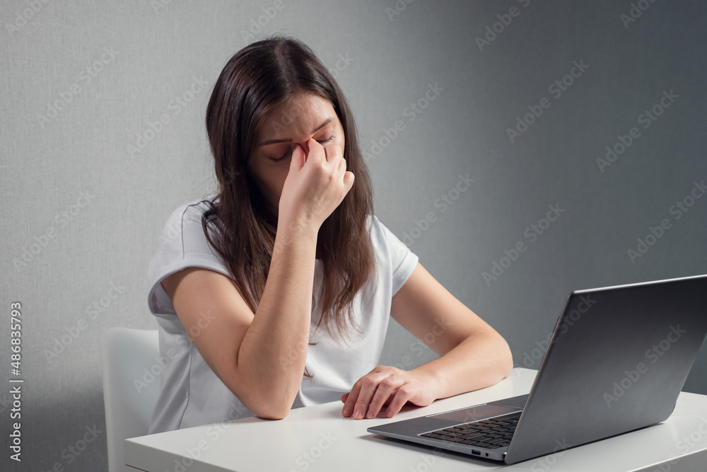 young woman massaging the bridge of nose and eyes while sitting in front of a laptop. young woman massaging the bridge of nose and eyes while sitting in front of a laptop.