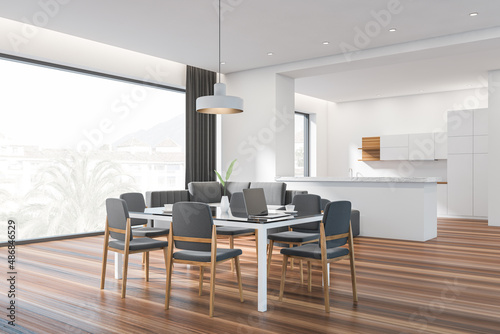 Modern dining room interior, panoramic window, large glass table with chairs. Stylish kitchen. hardwood flooring. Laptop. Couch. White open space area. 3d rendering.