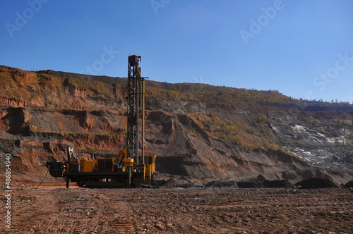 The drilling rig drills a hole for the subsequent laying of explosives into it. Industrial Blast Prep. In the background, an excavator is mining iron ore.