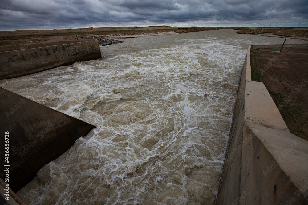 Fototapeta premium Aklak dam, Kzylorda region, Kazakhstan. Concrete channel on Shardara (Syrdarya) river. Turbulent yellow water, grey cloudy sky.