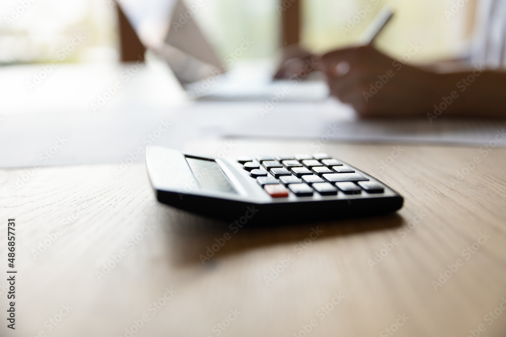 Close up focus on calculator with blurred young woman planning domestic monthly budget alone at home, managing household expenditures, doing financial accounting using e-banking computer application. Close up focus on calculator with blurred young woman planning domestic monthly budget alone at home, managing household expenditures, doing financial accounting using e-banking computer application.