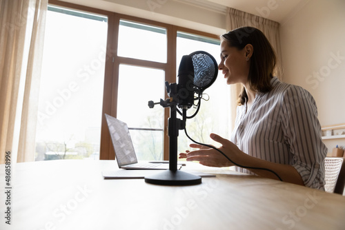 Fotografia, Obraz Smiling professional female radio host talking in stand microphone, looking at laptop screen, voice acting online, streaming live video lecture or educational webinar, vlogging at home office