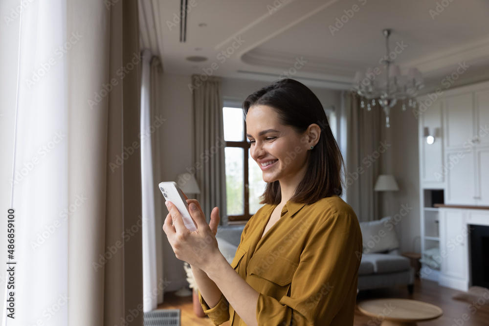 Side view happy beautiful young woman using cellphone applications, enjoying playing mobile games, web surfing, purchasing goods in internet store or communicating in social networks alone at home. Side view happy beautiful young woman using cellphone applications, enjoying playing mobile games, web surfing, purchasing goods in internet store or communicating in social networks alone at home.