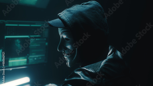 A masked hacker weared black hoodie uses a computer to stage a massive data attack on big data corporate servers, while sitting at desk in dark room and hacking database photo