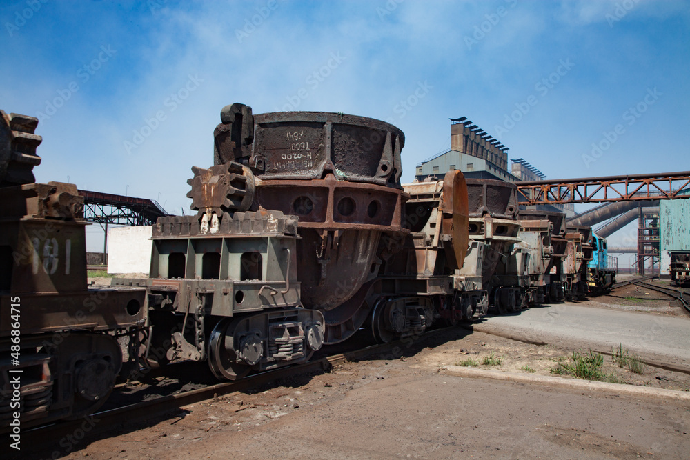 Metal alloys plant (smelter). Train on rails. Blue locomotive and rusted wagon. Metallurgical plant main industrial building on background.