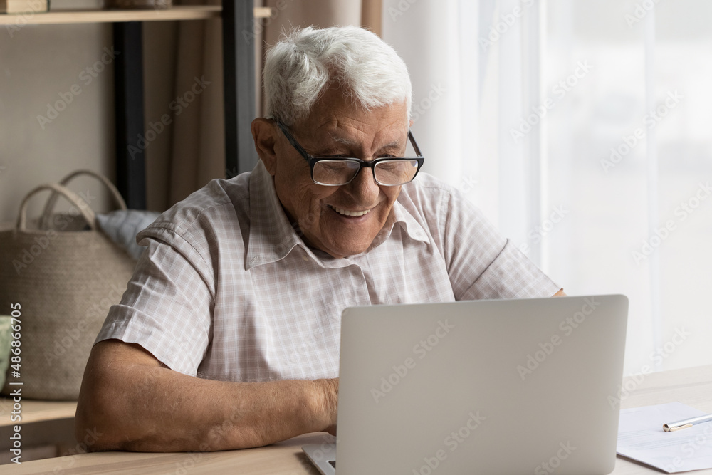 Happy senior older 80s man in glasses using laptop computer at work desk, typing, smiling, laughing, making video call, enjoying online talk, virtual chat, watching webinar, training on internet Happy senior older 80s man in glasses using laptop computer at work desk, typing, smiling, laughing, making video call, enjoying online talk, virtual chat, watching webinar, training on internet