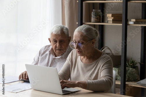 Senior married couple paying taxes, domestic bills, insurance fees on Internet. Husband and wife managing budget, counting expenses, savings, using laptop computer at table together
