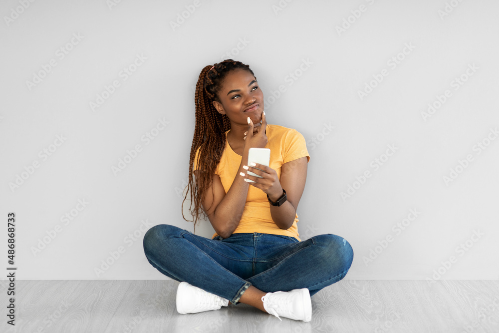 Thoughtful young black woman sitting cross legged on floor, using smartphone, thinking about something against grey wall Thoughtful young black woman sitting cross legged on floor, using smartphone, thinking about something against grey wall