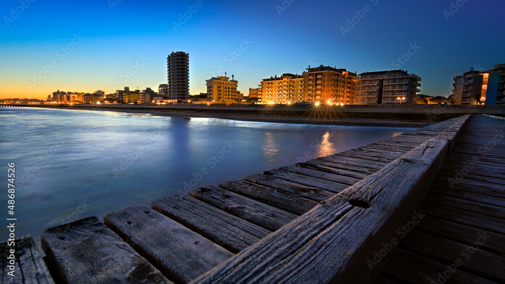 Jesolo seaside seen during the blue hour after sunset with blue sky and city lights seen from the wooden jetty on the beach.