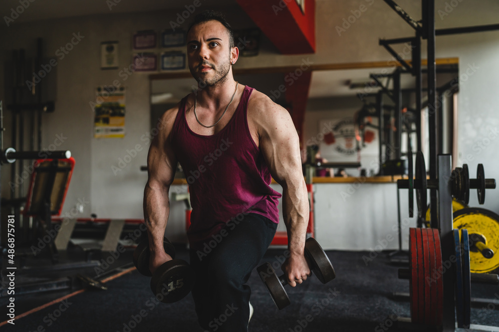 Fototapeta premium One man male bodybuilder training legs at the gym using dumbbells while doing lunges exercise real people copy space