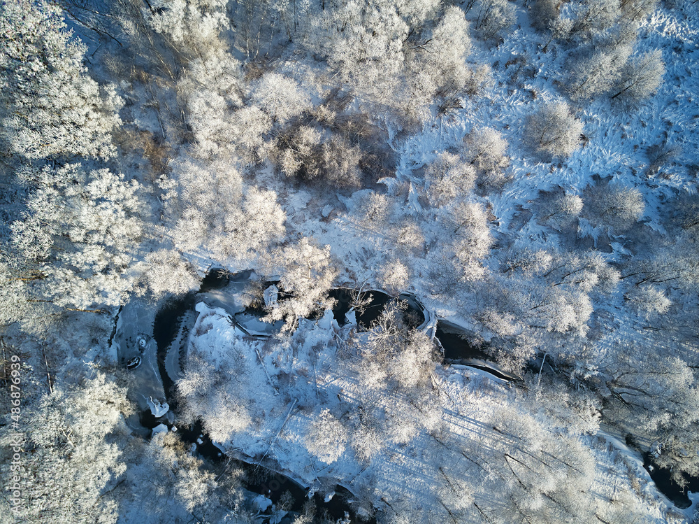 Winter woodland in frost top aerial view. Trees on riverbank. Small creek in forest. Rural dirt road. Snow covered field landscape.