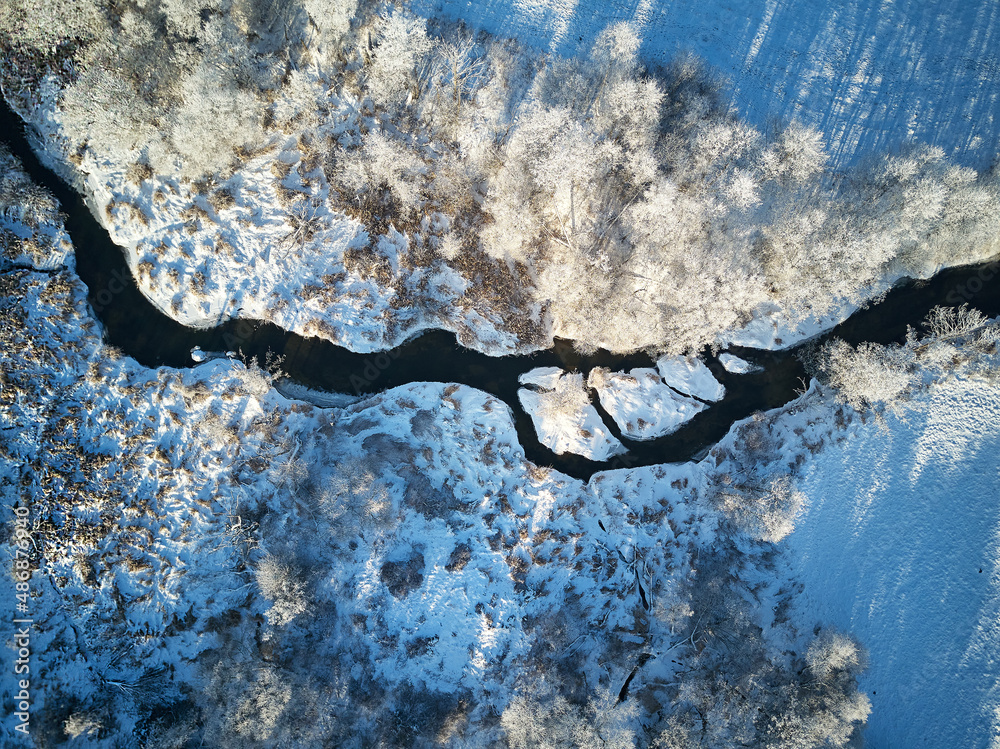 Winter woodland in frost top aerial view. Trees on riverbank. Small creek in forest. Rural dirt road. Snow covered field landscape. Cold weather