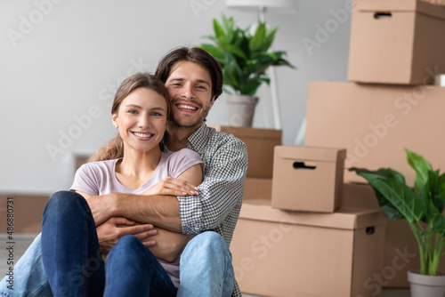Happy european millennial female and man in casual hugging sit on floor with cardboard boxes © Prostock-studio
