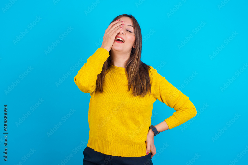 Young arab woman wearing knitted sweater over blue backgtound makes face palm and smiles broadly, giggles positively hears funny joke poses Young arab woman wearing knitted sweater over blue backgtound makes face palm and smiles broadly, giggles positively hears funny joke poses