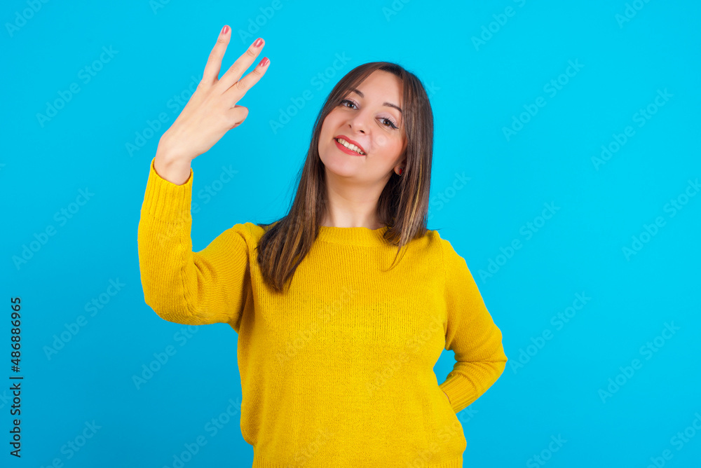 Young arab woman wearing knitted sweater over blue backgtound smiling and looking friendly, showing number three or third with hand forward, counting down Young arab woman wearing knitted sweater over blue backgtound smiling and looking friendly, showing number three or third with hand forward, counting down
