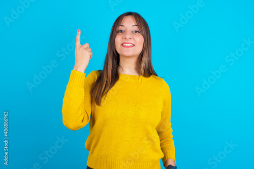 Young arab woman wearing knitted sweater over blue backgtound smiling and looking friendly, showing number one or first with hand forward, counting down © Roquillo