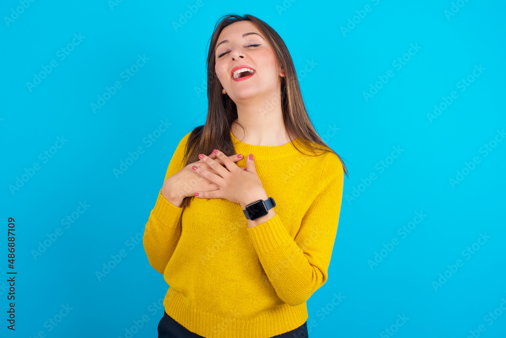 Young arab woman wearing knitted sweater over blue backgtound expresses happines, laughs pleasantly, keeps hands on heart