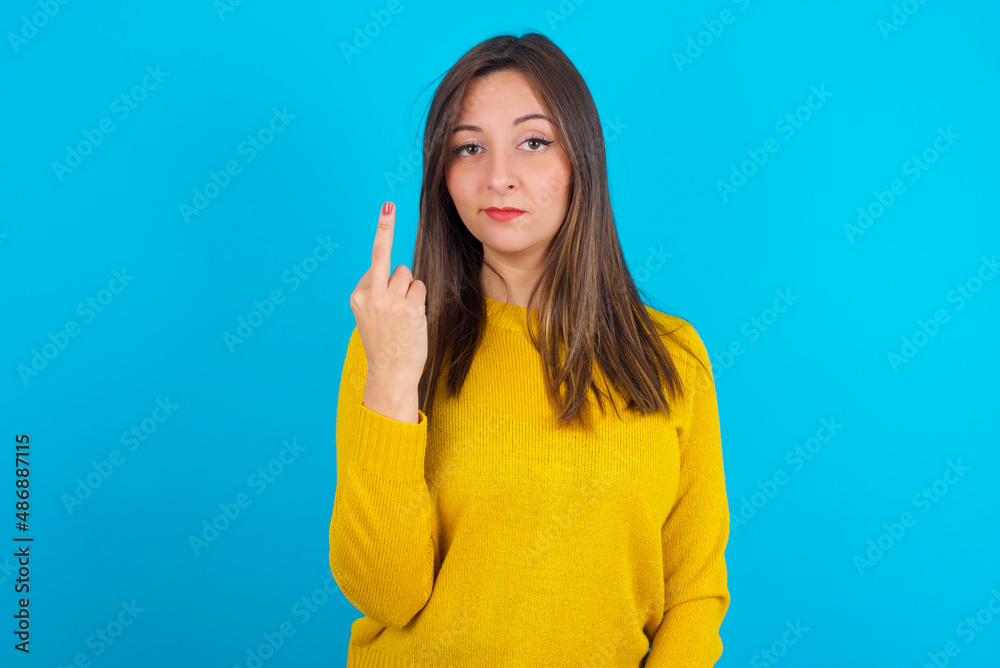 Young arab woman wearing knitted sweater over blue backgtound shows middle finger bad sign asks not to bother. Provocation and rude attitude. Young arab woman wearing knitted sweater over blue backgtound shows middle finger bad sign asks not to bother. Provocation and rude attitude.