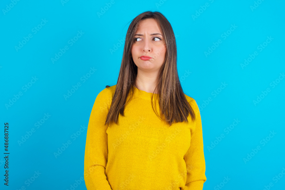 Dissatisfied Young arab woman wearing knitted sweater over blue backgtound purses lips and has unhappy expression looks away stands offended. Depressed frustrated model. Dissatisfied Young arab woman wearing knitted sweater over blue backgtound purses lips and has unhappy expression looks away stands offended. Depressed frustrated model.